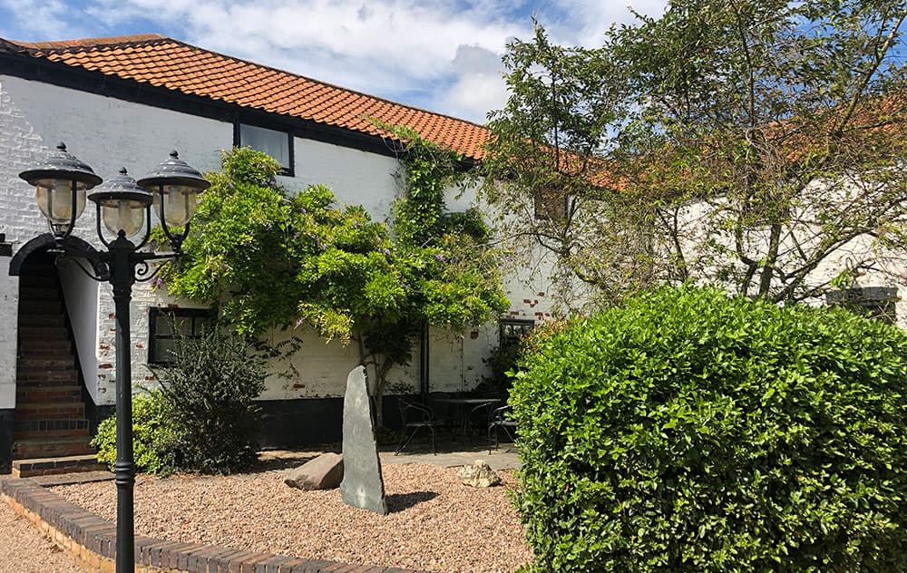 Courtyard garden with gravel, a standing stone, shrubs and a lamp post in front of whitewashed cottages with red tiled roofs