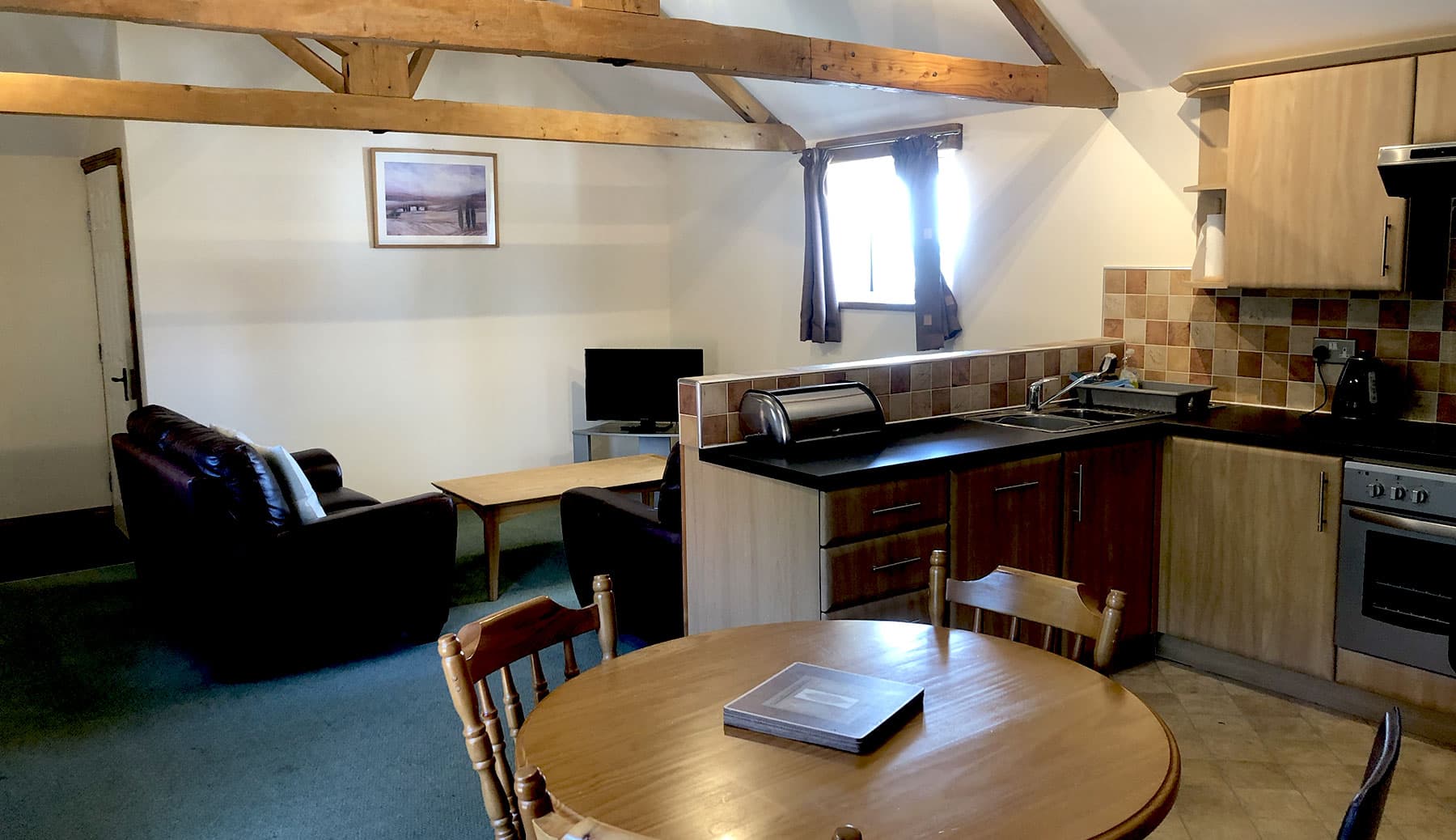 Open-plan room under exposed roof beams with a round dining table, a kitchen with oak units and a sitting area with a TV