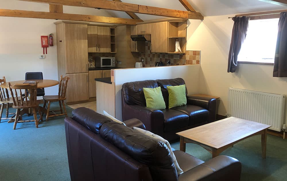 Open-plan living area with two leather sofas and a coffee table, with the kitchen and dining table behind, under exposed beams