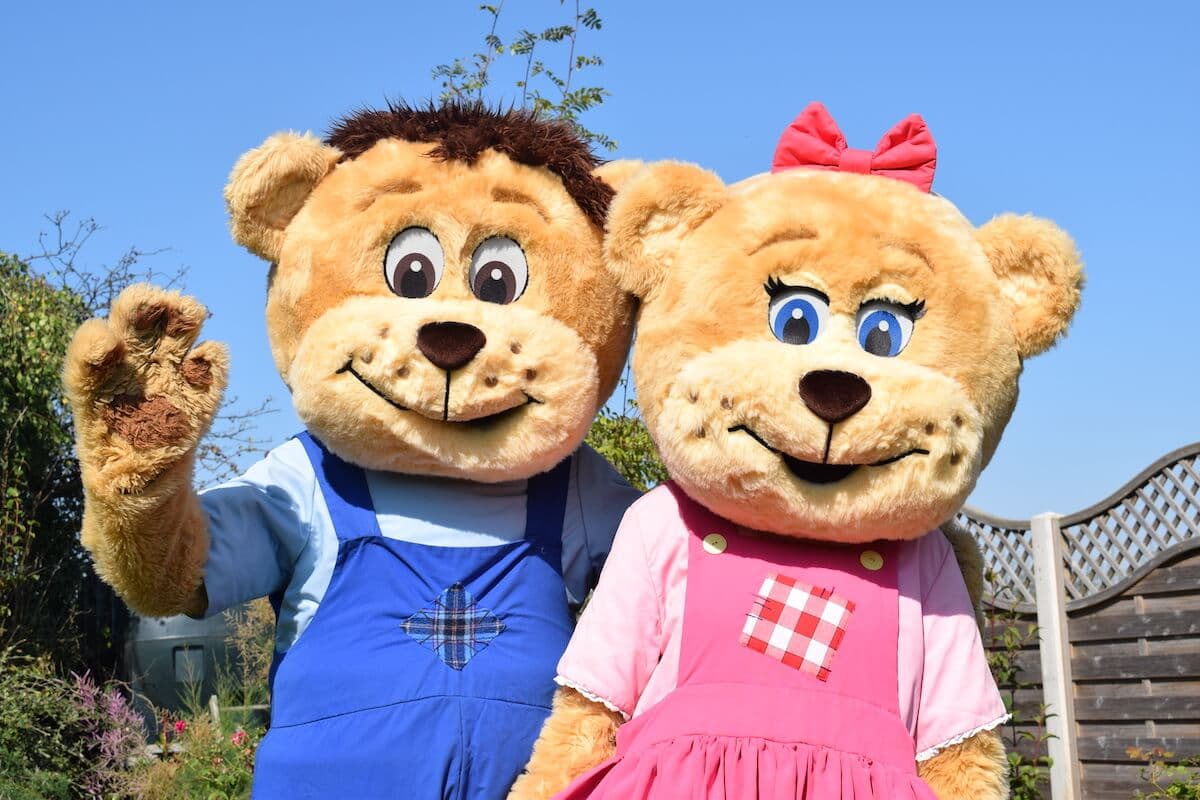 Two costumed bear characters, one in blue dungarees and one in a pink dress, waving in the sunshine