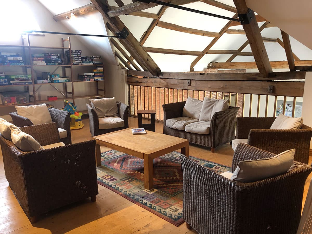 Seating area in a beamed loft with wicker armchairs and sofas round a coffee table on a rug, and shelves of board games