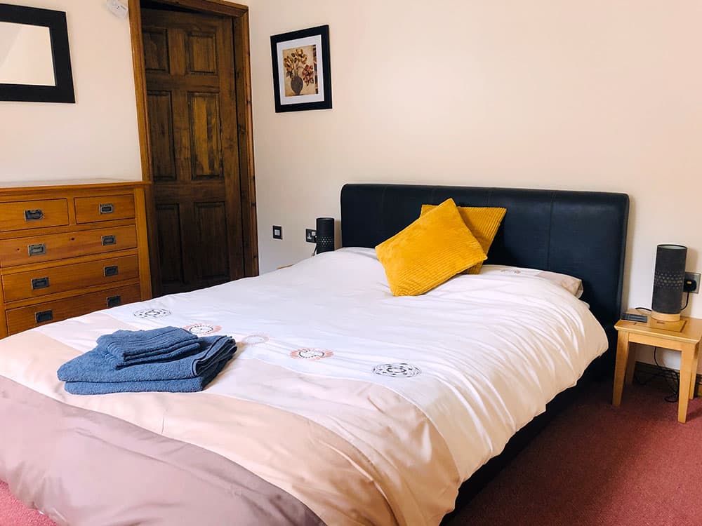 Double bed with a dark padded headboard, a yellow cushion and folded blue towels, beside an oak chest of drawers