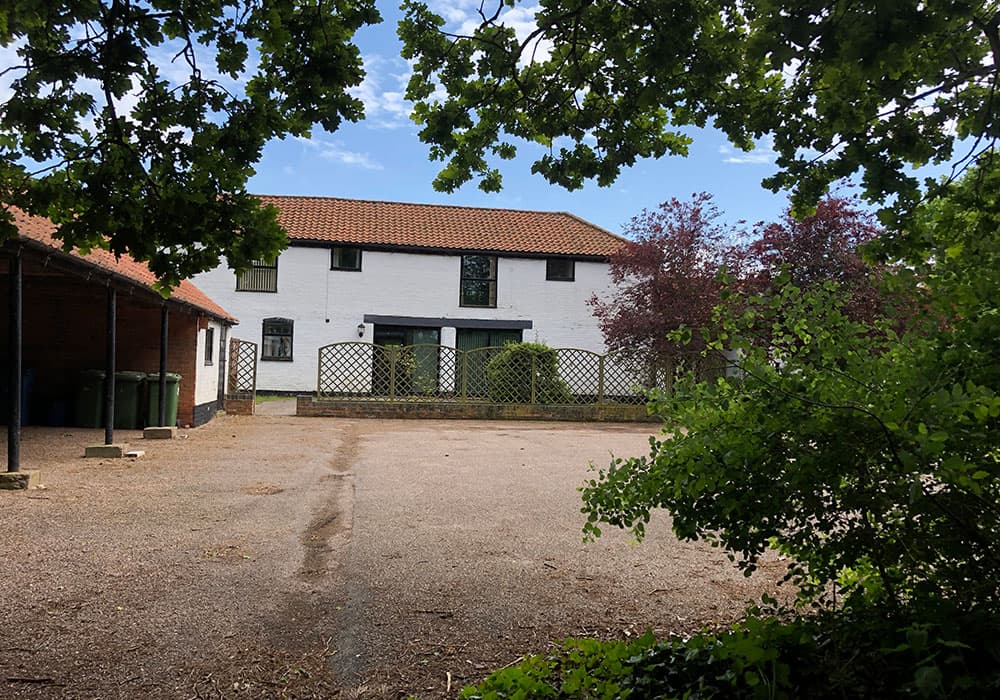 Two-storey whitewashed building with a red tiled roof, seen across a gravel yard framed by trees