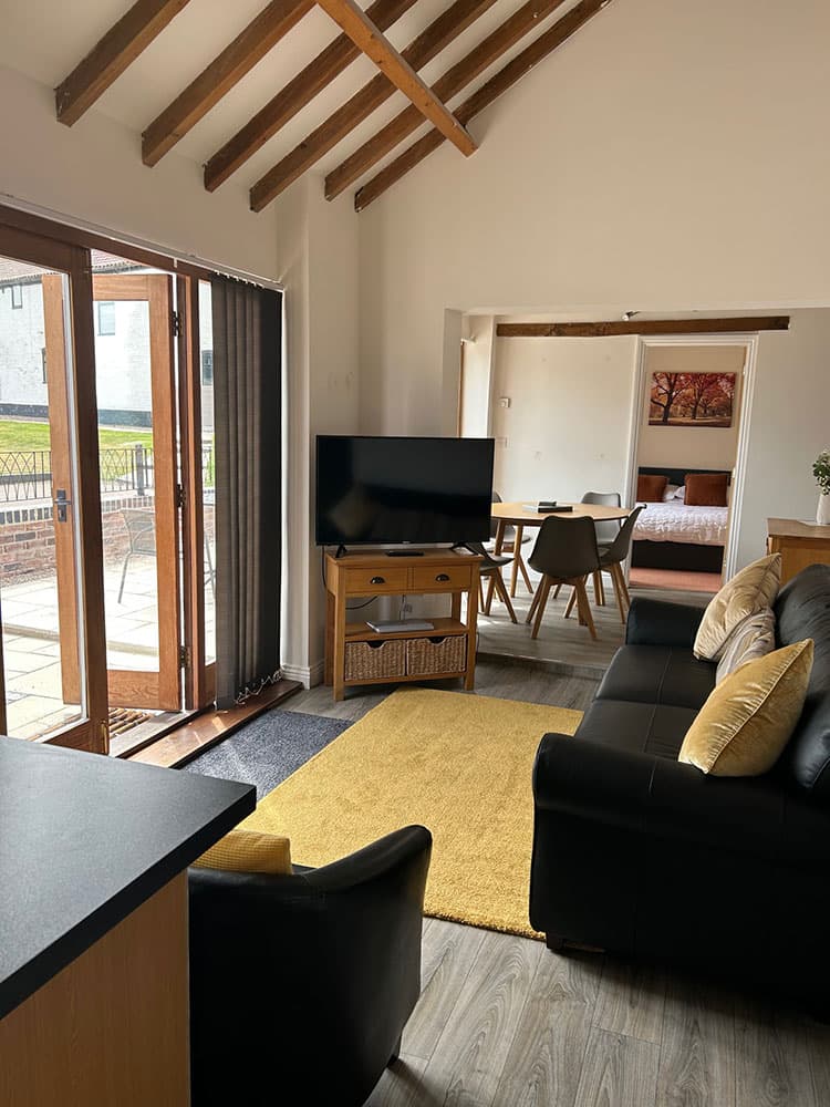 Open-plan living room under a beamed ceiling with black sofas, a yellow rug, a TV, glazed doors to the patio and a dining area beyond