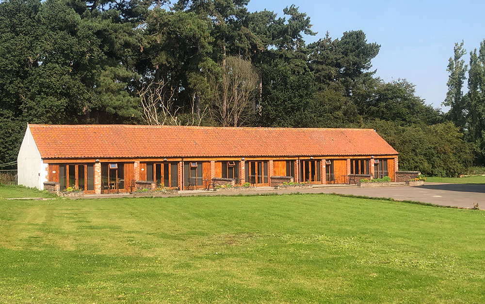Long single-storey brick building with a red tiled roof and glazed frontage, facing a large lawn with trees behind