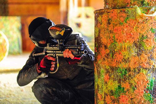 Paintball player in a mask taking aim from behind a paint-splattered barrel